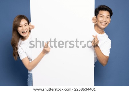 Young Asian couple is standing behind the white blank banner or empty copy space advertisement board and showing mini heart sign isolated on blue background, Looking at camera