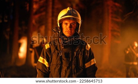 Portrait of a Confident Handsome Young Adult Firefighter in Safety Uniform and a Helmet with a Flashlight. Professional Fireman Looking at Camera. Wildland Fire in a Forest in the Background.