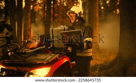 Brave Skillful Firefighter Standing Next to an ATV, Using Laptop Computer in Forest with Raging Brushfire. Superintendent or Squad Leader Making Sure Emergency Situation is Under Control. Royalty-Free Stock Photo #2258645735