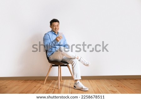 Excited middle aged asian man using smartphone while sitting on chair against white studio wall background, full length, free copy space Royalty-Free Stock Photo #2258678511