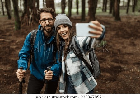 Cheerful young european guy and lady tourists in jackets with backpack and trekking sticks walk in forest, take selfie on phone, enjoy cold season outdoor. App for active lifestyle and travel blog