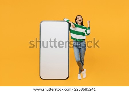 Full body young latin woman wear casual cozy green knitted sweater big huge blank screen mobile cell phone smartphone with area do winner gesture isolated on plain yellow background studio portrait