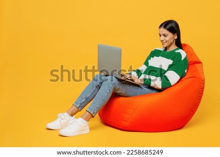 Full body young happy latin IT woman wears casual cozy green knitted sweater sit in bag chair hold use work on laptop pc computer isolated on plain yellow background studio People lifestyle concept