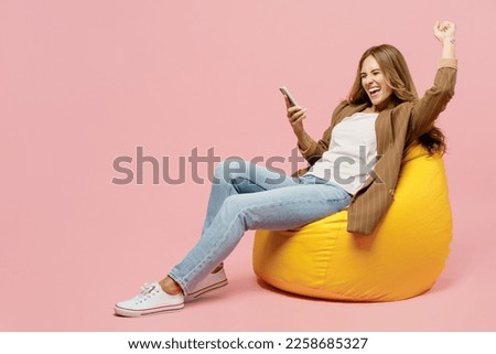 Full body young fun successful employee business woman 30s she wear casual classic jacket sit in bag chair hold use mobile cell phone do winner gesture isolated on plain pastel light pink background