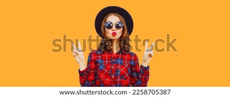 Portrait of stylish young woman blowing her lips sending kiss wearing black round hat and red shirt on orange background