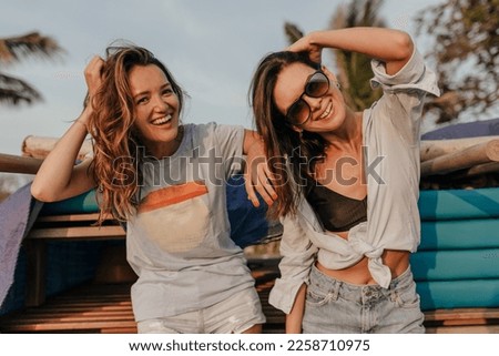 Two happy excited girls are laughing on shore of ocean on background go surfing boards on sunset. Outdoor photo of two female friends having fun on vacation