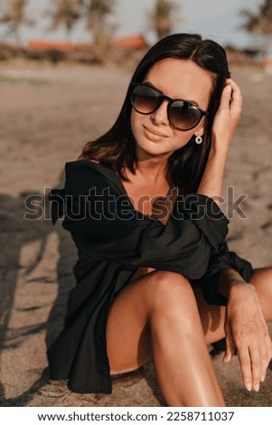 Stylish cute charming woman with dark hair wearing black shirt posing to camera with adorable smile and looks to camera on sandy beach
