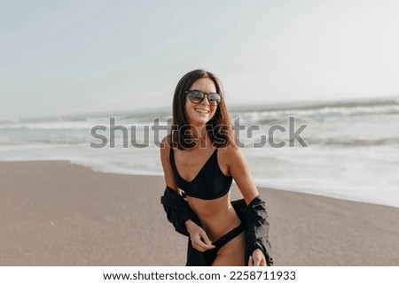 Cute attractive stylish lady with wonderful smile and dark hair wearing back shirt and swimsuit having fun, talking on the shore of ocean an enjoying vacation time.