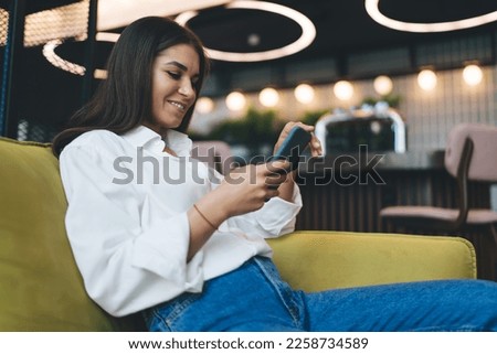Positive young female smiling and browsing while looking at screen of mobile phone and sitting in comfortable sofa during break in work against blurred cafe