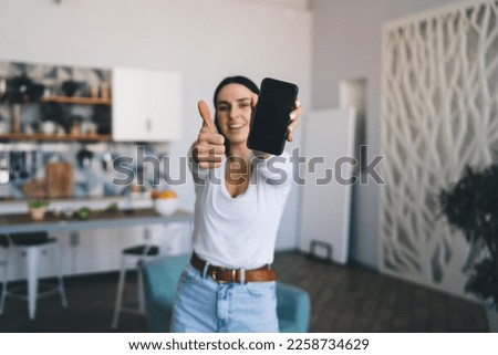 Selective focus of smartphone with black screen in hand of glad woman showing thumb up gesture while standing at home