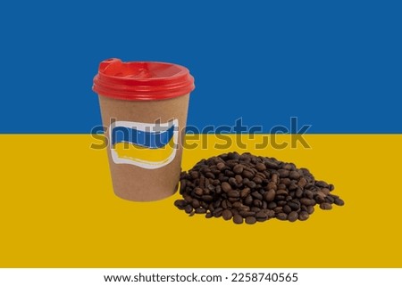 A disposable paper cup of coffee with a patriotic sticker and a handful of roasted coffee beans against the background of the national flag of Ukraine (close-up)