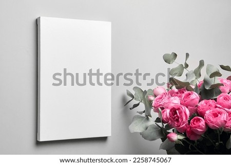 White canvas mockup hanging on grey wall and bouquet of pink roses with eucalyptus leaves. Empty blank canvas, interior decor Royalty-Free Stock Photo #2258745067