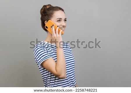 Side view of beautiful satisfied young adult woman wearing striped T-shirt talking via smart phone, having positive facial expression. Indoor studio shot isolated on gray background. Royalty-Free Stock Photo #2258748221