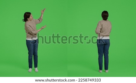 Casual smiling woman pointing at presentation icon in studio, doing advertisement and showing example on camera. Person posing over isolated greenscreen backdrop, confident girl doing ad.