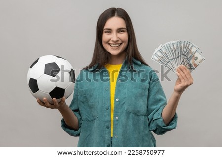 Portrait of smiling woman showing soccer ball and fun of hundred dollar bills, winning lot of money betting for sport, wearing casual style jacket. Indoor studio shot isolated on gray background.