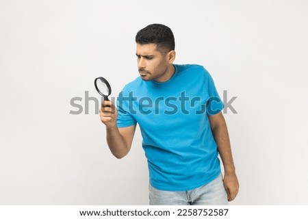Portrait of unshaven man wearing blue T- shirt standing looking through magnifying glass, finding out something, exploring, inspecting. Indoor studio shot isolated on gray background.