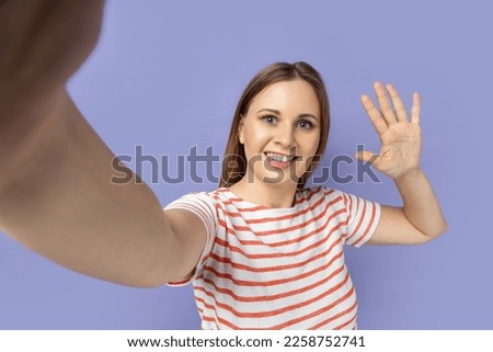 Portrait of attractive woman taking selfie picture, point of view, looking at the camera and waving hand, saying hi, making front selfportrait. Indoor studio shot isolated on purple background.