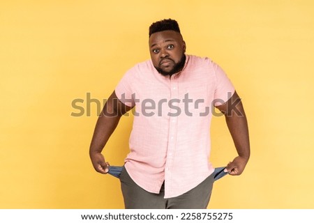 Portrait of frustrated worried man wearing pink shirt turning out empty pockets showing no money gesture, frowning face. Indoor studio shot isolated on yellow background. Royalty-Free Stock Photo #2258755275