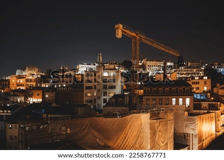 A top-down view of a city under construction at night in low-key and long exposure. Old residential houses surround the on-site building, a tall yellow crane towering over the buildings standing out Royalty-Free Stock Photo #2258767771