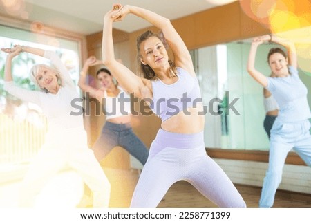Class of cheerful young old female dancers raising both hands above their heads while exercising in room of wellbeing fitness center
