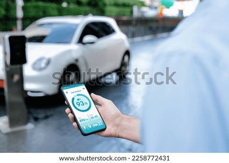 Closeup progressive businessman look at EV car's battery status application on smart phone screen at public parking charging station with power cable plug and renewable energy-powered electric vehicle
