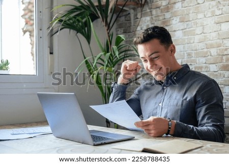 Excited business man student reading postal mail letter overjoyed by great news, happy male winner holding paper bill with loan approval celebrate taxes refund receive salary rise payment sit at desk Royalty-Free Stock Photo #2258788435