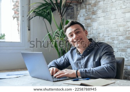 Good-looking millennial office employee in glasses sitting at desk in front of laptop smiling looking at camera. Successful worker, career advance and opportunity, owner of prosperous business concept Royalty-Free Stock Photo #2258788945