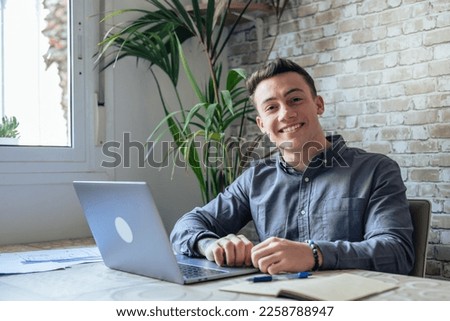 Good-looking millennial office employee in glasses sitting at desk in front of laptop smiling looking at camera. Successful worker, career advance and opportunity, owner of prosperous business concept Royalty-Free Stock Photo #2258788947