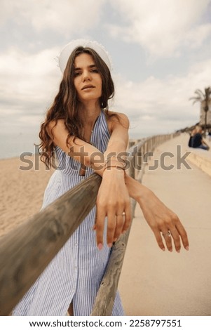 Cute young caucasian girl with wavy hair leaning on railing looking at camera. Brunette model wears sun-dress stands on coast. Concept seaside vacation.