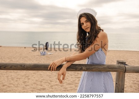Beautiful young caucasian woman stands on beach sand with sea in background. Brunette wears sun-dress and cap looking at camera outside. Concept seaside vacation.