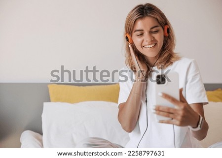 Young caucasian girl speaking talking on video call with smartphone and headphones indoor. Blonde hair woman in white t-shirt sitting on bed. Concept communication, calls.