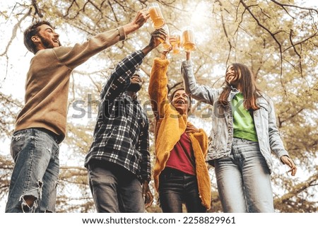 A group of four young adults standing and toasting with beer glasses raised high. Filled with joy and excitement as they celebrate in the beautiful countryside, greenery and bright sunshine