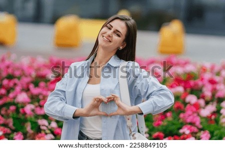 Excited caucasian brunette girl in casual smiles looks at camera makes heart gesture standing against blurry flowers at shopping mall yard. American young woman in love expressing feelings, romance.