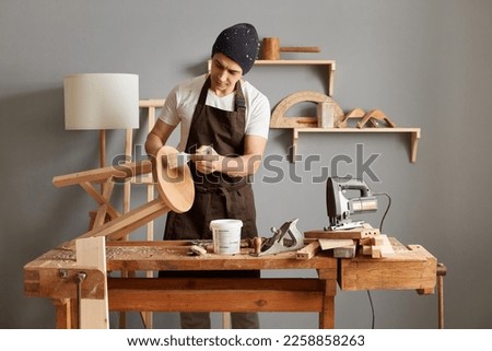 Craftsman and DIY handmade. Image of handsome man carpenter wearing brown apron and black cap applies paint using paintbrush in carpentry workshop,painting furniture with a paintbrush. Royalty-Free Stock Photo #2258858263