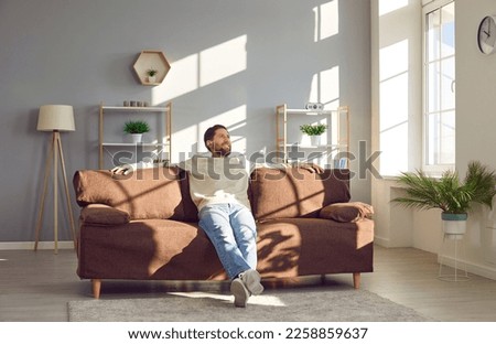 Man relaxing on the sofa at home. Happy young man sitting on a comfortable brown couch in a spacious living room interior with modern furniture, house plants and big windows Royalty-Free Stock Photo #2258859637