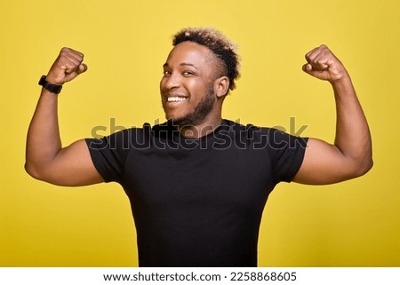 An African-American smiling male athlete in a black T-shirt rejoices in victory on a yellow background. A confident, happy man demonstrates biceps, muscles or strength and looks at the camera