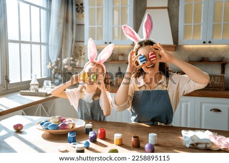 Happy mother and her daughter wearing aprons holding painted colorful eggs while decorating them with food dyes in cozy kitchen at home. Easter craft activities for families.
