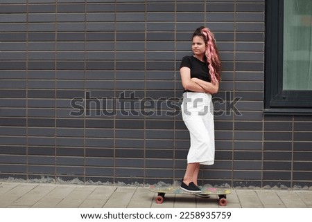 Sad teenage girl with skateboard stands full length on street next to wall of gray brick. Crisis of adolescence. Authentic lifestyle portrait of teenager