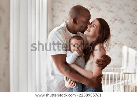 Happy multiethnic family hugging at home. Mother and father kiss while holding child in arms, who looks camera Royalty-Free Stock Photo #2258935761