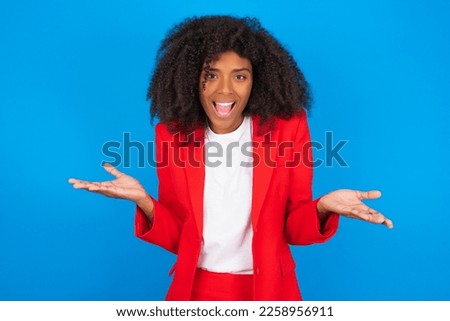 So what? Portrait of arrogant young businesswoman with afro hairstyle wearing red over blue wall shrugging hands sideways smiling gasping indifferent, telling something obvious. Royalty-Free Stock Photo #2258956911