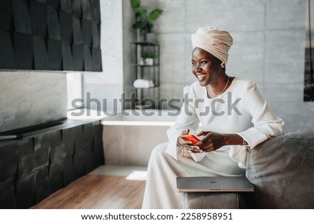 Happy African woman in white dress and turban holds phone sitting at home on couch. Happy African American businesswoman laughs looks away being satisfied. People candid emotions. Woman in hotel. Royalty-Free Stock Photo #2258958951