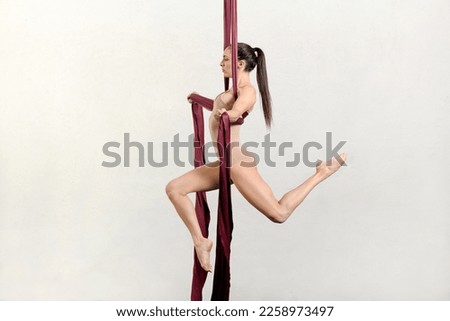 Side view of alluring young fit female dancer with long dark hair in beige bodysuit performing cross pose on aerial silks against white background