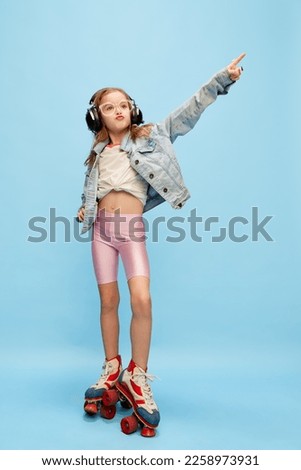 Music. Little beautiful girl, child in shorts and jeans jacket posing in headphones on rollers over blue studio background. Concept of childhood, emotions, fun, fashion, lifestyle, facial expression Royalty-Free Stock Photo #2258973931