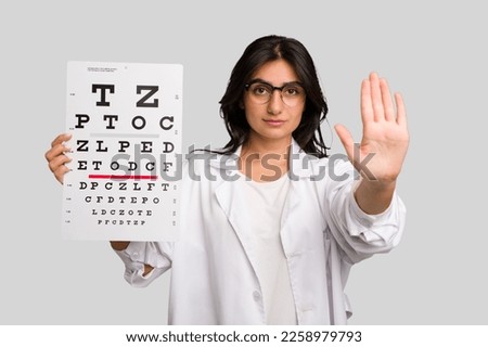 Young indian oculist woman holding an eye chart paper cut out isolated standing with outstretched hand showing stop sign, preventing you.