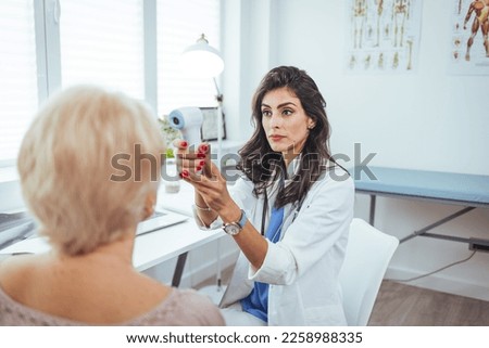 Young nurse or doctor taking patient's body temperature with non-contact infrared thermometer. Woman gets temperature measured during visit to hospital for flu or Covid-19 vaccine