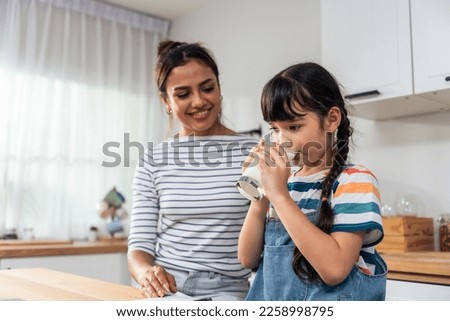 Caucasian little kid holding a cup of milk and drinking with mother. Attractive mom teach and support young girl daughter take care of her body, sipping a milk after wake up for health care in house. Royalty-Free Stock Photo #2258998795