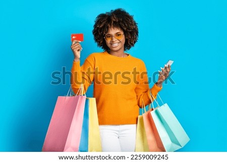 Cool stylish happy smiling young black woman with bushy hair wearing sunglasses holding colorful shopping bags, smartphone, credit card, posing over blue studio background. E-commerce, retail concept