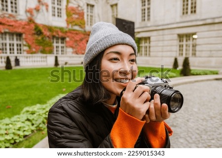 Beautiful young smiling asian woman in warm clothes taking pictures with vintage camera while sitting in old city