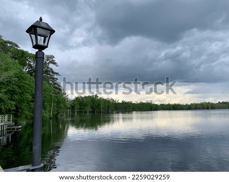 Look down the still, clam water of the Hope Mills lake. Dense forest surrounds the lake and an antique, traditional looking lamppost provides light.