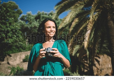 Cheerful Hispanic female smiling and looking away while standing in green park and taking photos on photo camera in bright sunlight during vacation